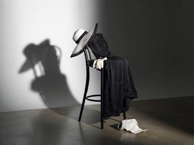 A wide-brimmed hat, white gloves, and a sparkly black dress are arranged on a tall black wooden chair next to a clutch purse and paper on the floor.