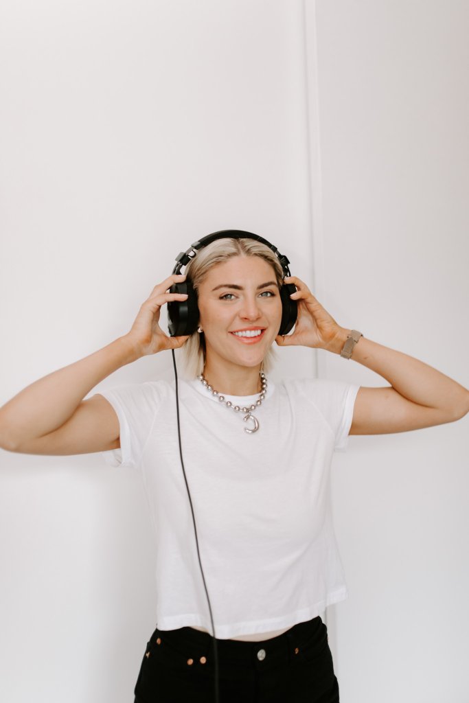 Courtney Johnson wearing wired headphones over a white t-shirt, smiling at the camera against a white wall
