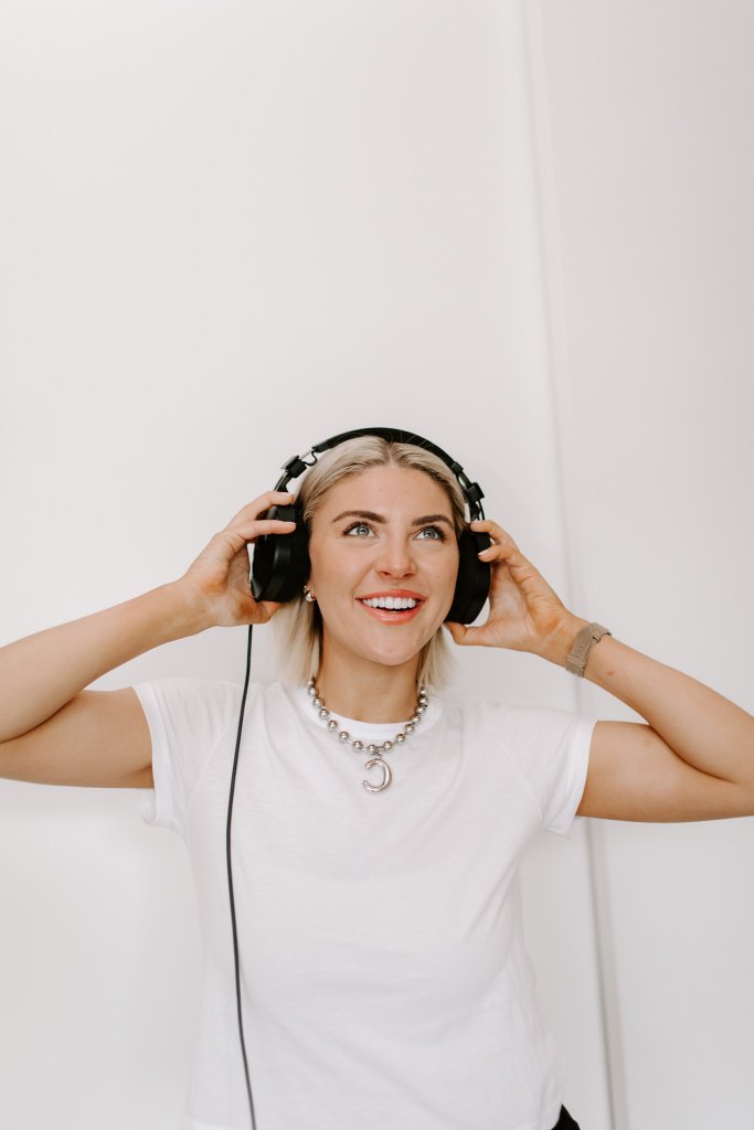 Courtney Johnson in a white t-shirt wearing wired headphones, looking up with an open smile against a white wall